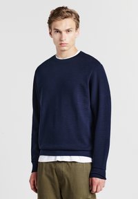 Felpa di un blu navy dal tessuto texturizzato con maniche lunghe e polsini a costine, indossata sopra una maglietta bianca a collo tondo, abbinata a pantaloni verde oliva.
