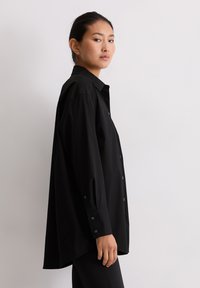 Vrouw die zijwaarts staat, gekleed in een losvallend, lang zwart overhemd met knopen en zwarte broek, tegen een effen lichte achtergrond.