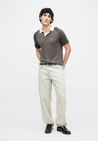 Polo marron à manches courtes avec un col crème, associé à un jean large beige clair et des chaussures noires, avec un branding minimal.