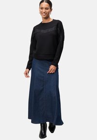 Černá mikina se třásněmi na hrudníku, kombinována s dlouhou džínovou sukní. Modelka má na sobě černé kotníkové boty.