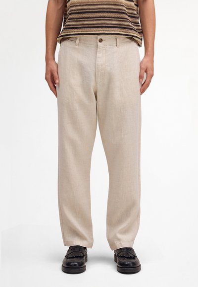 Mann trägt beige Leinenhosen mit Knopf und Gürtelschlaufen, kombiniert mit schwarzen Loafers, steht vor einem schlichten weißen Hintergrund.