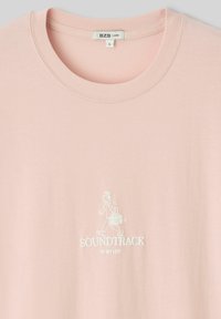 T-shirt rose clair avec un col rond, présentant un graphique brodé blanc d'une personne marchant avec un boombox et le texte "SOUNDTRACK OF MY LIFE".