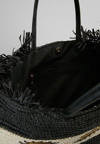 Sac cabas tissé noir et naturel avec paille texturée, bords frangés, poignées élégantes et poche intérieure zippée. Fermeture à pression bronze.