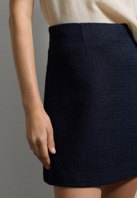 Gonna mini in tweed blu navy con un aspetto testurizzato e vita aderente, abbinata a un top in un colore neutro, con la mano appoggiata sul tessuto.