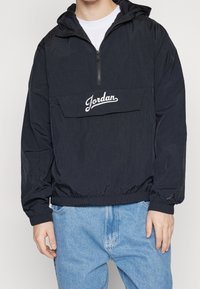 Black pullover jacket with a zipper, hood, and elastic cuffs. Features a front pocket with the "Jordan" logo. Made from lightweight material.