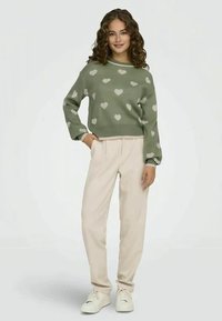 Žalios megztos džemperis su švelnių širdžių raštais, apvalia iškirpte ir ilgomis rankovėmis, derinamas su béžinėmis kelnėmis ir baltomis sportinėmis bateliais.