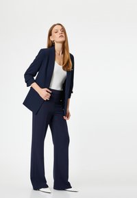Blazer azul marino de corte entallado, acompañado de unos pantalones de pierna ancha a juego. Una blusa blanca y unos zapatos blancos de punta completan el conjunto.