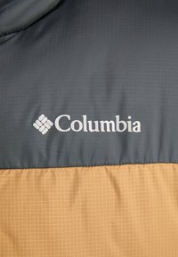 Doudoune avec une partie supérieure gris foncé et un bas à motif carreaux brun clair. Logo "Columbia" imprimé en blanc sur la section supérieure.