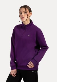 Sudadera de felpa morada con un alto cuello, cremallera frontal de media longitud, puños acanalados y un pequeño logo en el pecho. Textura suave.