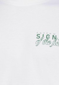 Witte katoenen t-shirt met groene tekst die leest "volg de tekenen van de toekomst", gecombineerd met vetgedrukte en cursieve typografie.