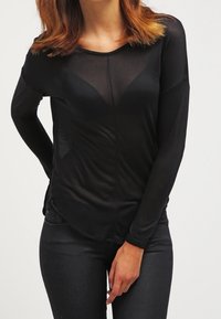 Žena v černém průhledném topu s dlouhými rukávy přes černou podprsenku, v černých kalhotách, s jednou rukou opřenou o bok a druhou před sebou.