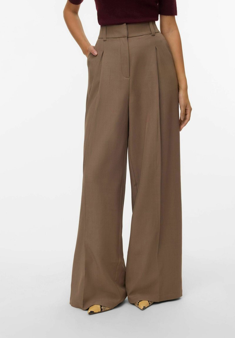 Pantalones marrones de talle alto y pierna ancha, hechos de un material suave, con pliegues frontales y detalles ajustados. Combinando con calzado estampado.