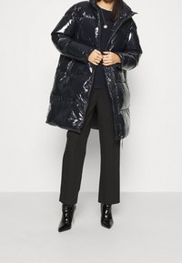 Schwarze, glänzende Steppjacke mit hohem Kragen, Reißverschluss vorne und Seitentaschen. Kombiniert mit schwarzen Hosen und glänzenden Stiefeletten.