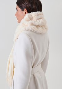 Femme portant un manteau blanc ceinturé et une écharpe crémeuse et fluffy, vue de côté sur un fond uni.