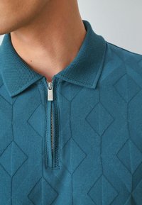 Tyrkysová polo košile se strukturovaným diamantovým vzorem, s límcem na zip a stříbrnými detaily. Měkká pletená tkanina se strukturovaným designem.