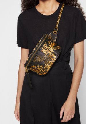 Kobieta ubrana w czarny strój, z czarno-złotą torebką na pasku Versace Couture przewieszoną przez pierś, z złotym łańcuszkiem i zamkiem błyskawicznym.