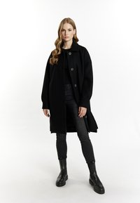 Kvinde med langt bølget hår, der står iført en sort knaplukket frakke, sort top, sorte slim-fit jeans og sorte chunky støvler foran en hvid baggrund.