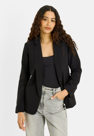 Joven mujer con cabello largo y rizado, vistiendo una chaqueta negra sobre una camiseta negra y vaqueros gris claro, de pie con una mano en el bolsillo.