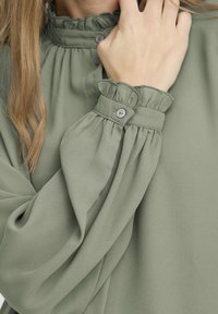 Green blouse with ruffled neckline and cuffs, featuring button closures. Fabric appears soft and drapes gently.