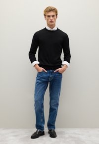 Schwarzer Pullover mit langen Ärmeln, blaue Denim-Jeans und schwarze Schuhe. Weißer Kragen des Hemdes sichtbar. Model posiert mit den Händen in den Taschen.