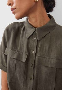 Kurzes, olivgrünes Leinenhemd mit Kragen, zwei aufgesetzten Taschen und Knopfverschluss. Weiche Textur und glatte Stoffoberfläche.