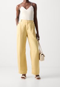 White ribbed tank top with chain straps, paired with loose yellow trousers and white heeled sandals. Accessorized with a cream handbag.