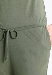 Tissu vert olive avec une texture douce, doté d'un cordon de serrage noué à la taille et de poches latérales, mettant en avant un design à coupe décontractée.