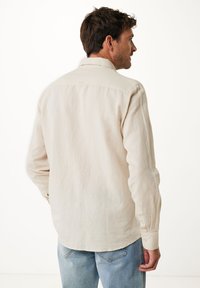 Chemise beige à manches longues, en tissu léger, avec col, poignets boutonnés et ourlet courbé. Portée avec un jean bleu clair.