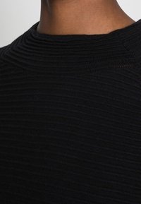 Svart stickad topp med en texturerad yta. Hög, rund krage utan utsmyckningar, som visar en ribbad struktur genom hela plagget.