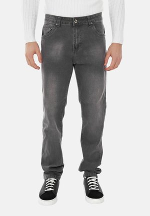 Sneakers nere con lacci bianchi, jeans grigi sbiaditi e una maglia a maniche lunghe bianca a coste indossati da una persona in piedi, vista frontale, isolata su sfondo bianco.