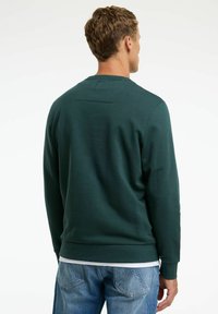 Hanorac verde închis cu un design texturat, manșete și tiv côté, având un mic accent rectangular pe spatele superior. Aproape cu blugi.