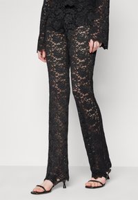 Svarta spetsbyxor med ett blommönster, utsvängd design och genomskinlig textur, tillsammans med svarta sandaler med remmar.