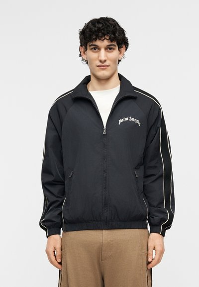 Noor tumeda lokkis juustega mees kannab musta tõmblukuga jopet valge ääristusega ja rinnal on kiri "Palm Angels", mida ta kannab beežide pükstega.