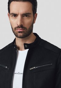 Czarna kurtka z wysokim kołnierzem, kieszeniami na zamek i teksturowanym materiałem. Noszona na białym T-shircie z nadrukowanym tekstem.