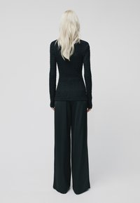 Una persona bionda sta di spalle, indossando un maglione lungo aderente nero a trama e pantaloni neri larghi, su uno sfondo semplice.