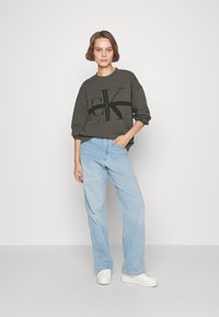 Sudadera gris con gran logo negro 'CK', combinada con jeans de pierna ancha de color azul claro. Modelo usando zapatillas blancas, de pie frente a un fondo liso.