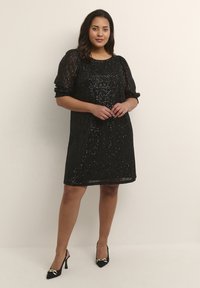 Robe noire à sequins avec des manches bouffantes en tulle, décolleté en scoop et coupe ample. Associée à des talons noirs pointus avec des accents dorés.