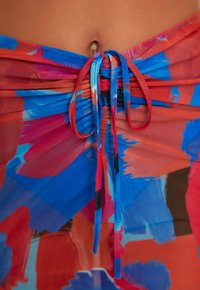 Partie médiane d'une personne avec un tissu coloré bleu et rouge à motifs noué en nœud autour de la taille, exposant un nombril.