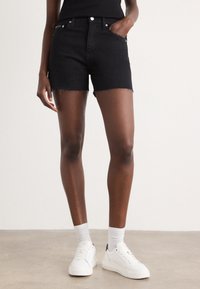 Black denim shorts with frayed hem, designed with a five-pocket style and metal hardware, paired with white sneakers and socks.