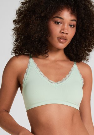 Vrouw met krullend haar die een lichtgroene, geribbelde bralette met kanten rand draagt, poserend tegen een effen grijze achtergrond.