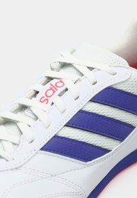 Zapatilla deportiva blanca con rayas moradas, paneles de malla texturizada y cordones planos. Destaca el logotipo rojo en la lengüeta y una suela acolchada.