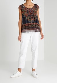 Sort blomsterbluse med korte, gennemsigtige ærmer, rynket halsudskæring og indviklede mønstre. Kombineret med hvide cropped bukser og hvide sneakers.