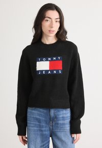 Svart strikket genser med ribbede detaljer, med et fremtredende logo-patch i rødt, hvitt og blått. Brukt med lyseblå jeans.