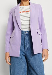 Blazer lavanda con solapas puntiagudas y dos bolsillos frontales, combinado con una camisa a rayas y pantalones vaqueros de pierna ancha de cintura alta.