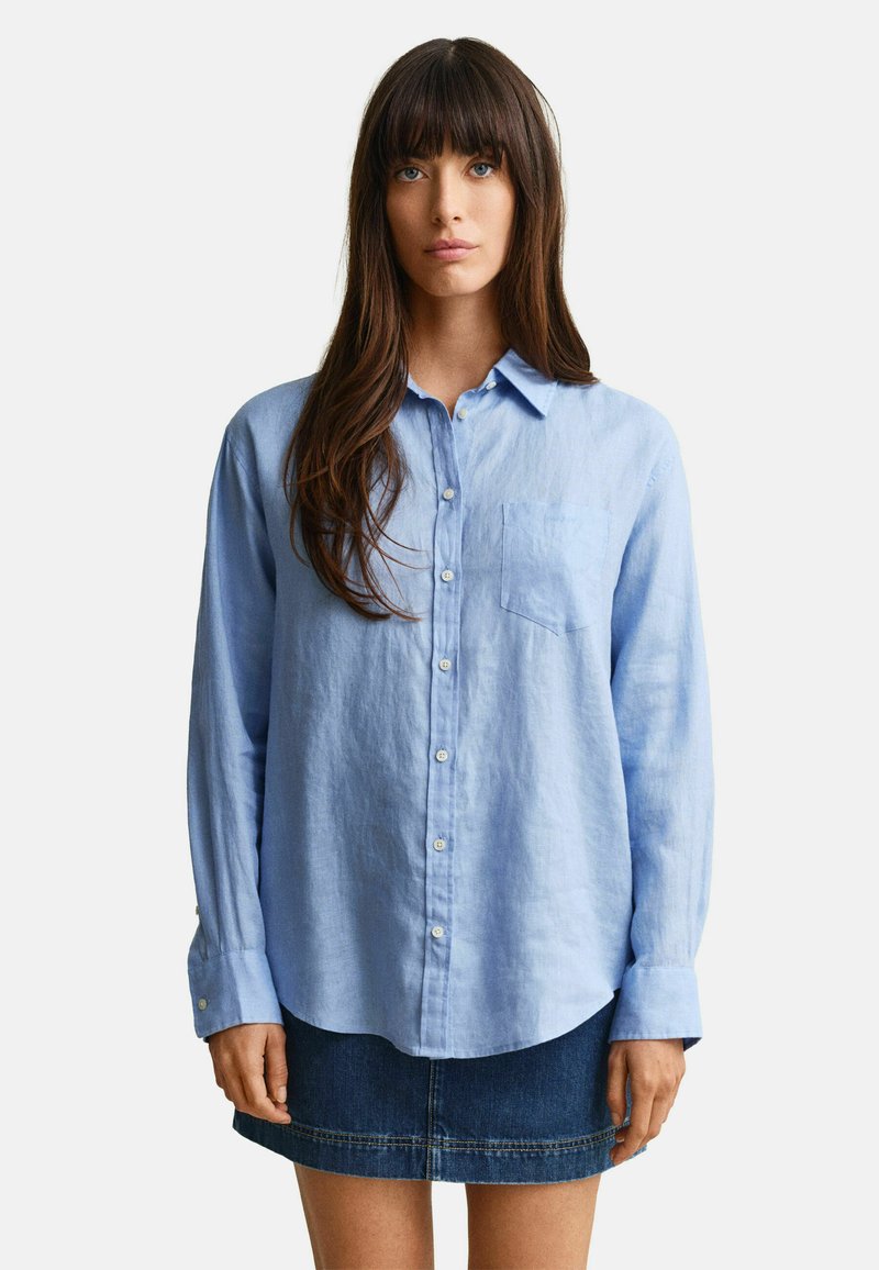Femme aux cheveux longs et bruns portant une chemise bleu clair à boutons et une jupe en jean foncé, debout devant un fond blanc uni.