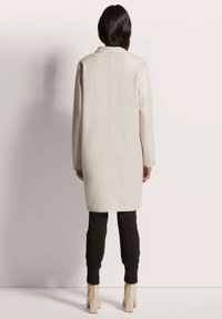 Hellbeige langer Mantel aus strukturiertem Stoff, lässige Passform, Kragen und gerade Mittelnaht, kombiniert mit schwarzen Hosen und cremefarbenen Knöchelstiefeln.