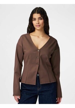 Kobieta z długimi kręconymi włosami nosi brązowy rozpinany sweter z dekoltem w serek oraz ciemnoniebieskie dżinsy, stoi na tle jednolitego jasnego tła.