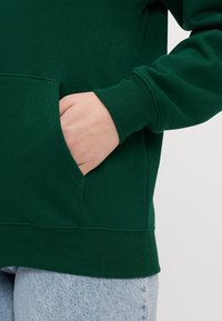 Vihreä huppari, jossa on etutasku, ribbireunat ja helma, valmistettu pehmeästä kankaasta. Taskussa näkyy käsi. Myös vaalean siniset farkut näkyvät.