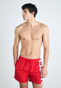 Shorts de bain rouges avec une taille élastique et un cordon de serrage ; présente un grand logo blanc sur le côté ; fabriqués en matériau léger et à séchage rapide.