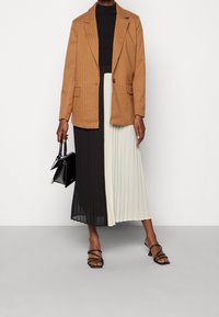 Blazer marrone a righe sottili su una gonna plissettata bianca e nera. Accessoriato con una piccola borsa nera e sandali neri con cinturini.